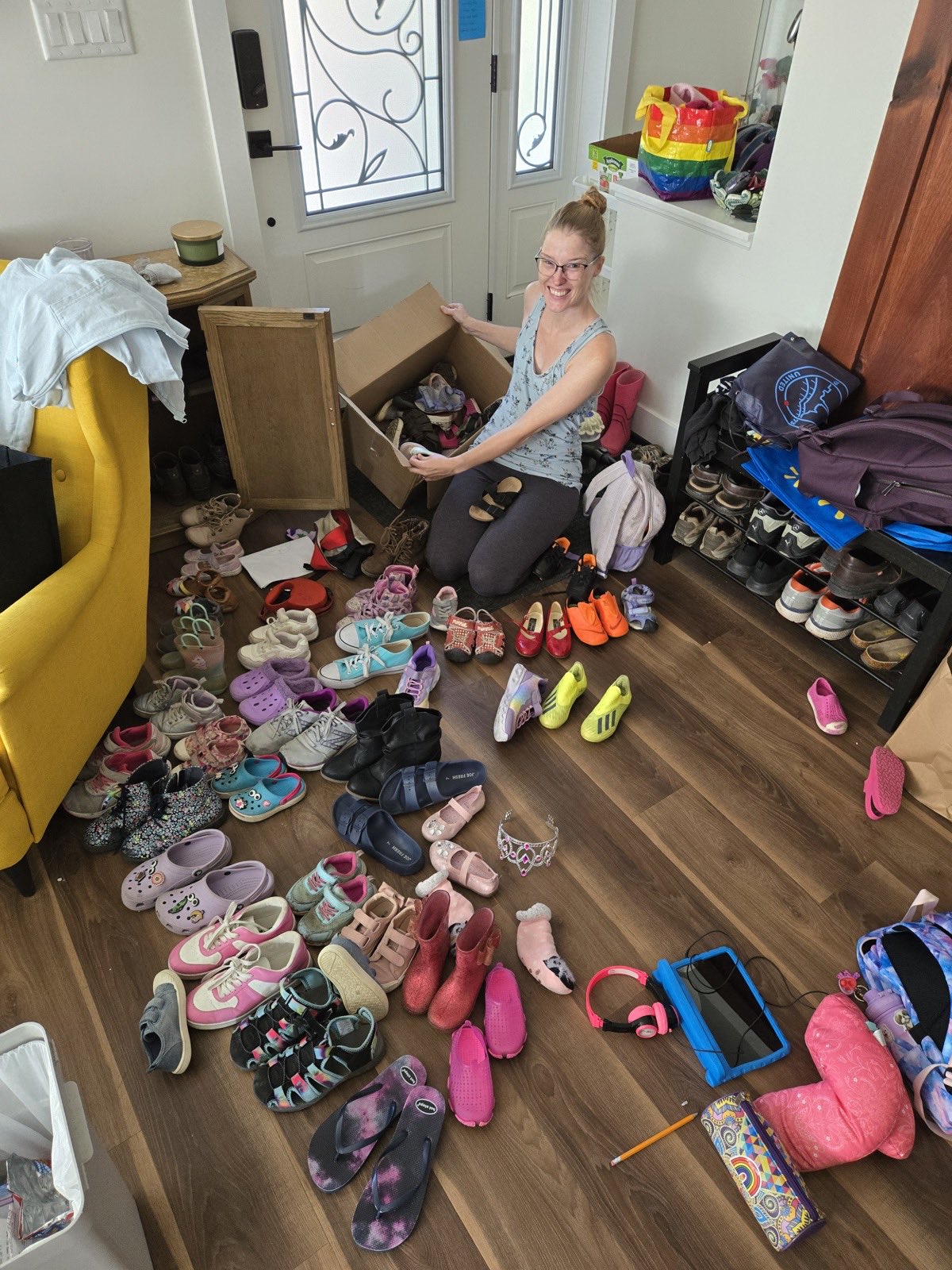 Megan kneeling on a hardwood entryway floor beside an open cardboard box, sorting dozens of pairs of children's shoes and boots spread out around her.