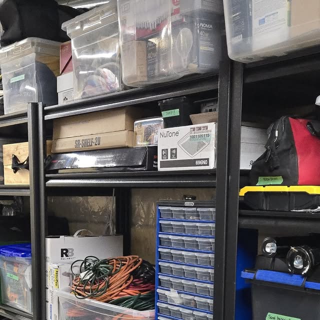 Garage utility shelving with clear labelled bins, a blue multi-drawer parts organizer, coiled extension cords and stacked boxes arranged on black metal racking.
