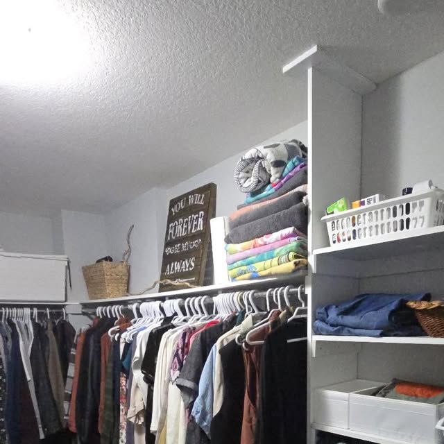 Walk-in closet with clothing hung on matching white hangers, folded stacks and labelled white baskets on the shelf above, and a wooden "You will forever be my always" sign leaning at the back.