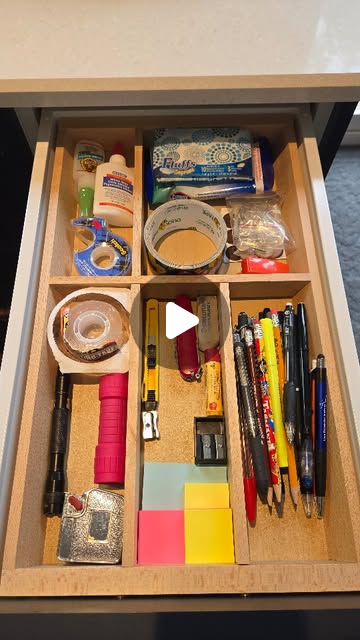 Open drawer fitted with a wooden divider tray, sorting pens, pencils and markers, rolls of tape, glue, a utility knife and coloured sticky notes into separate compartments.