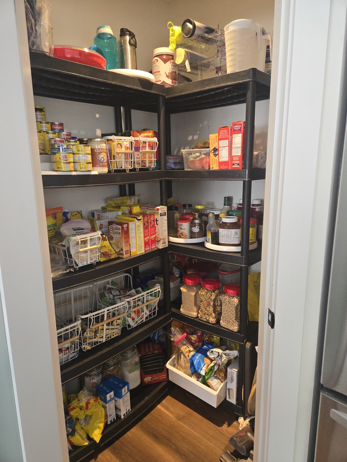 Corner pantry with dark wire shelving, canned goods and boxed food grouped by type, and dry goods decanted into labelled clear jars.