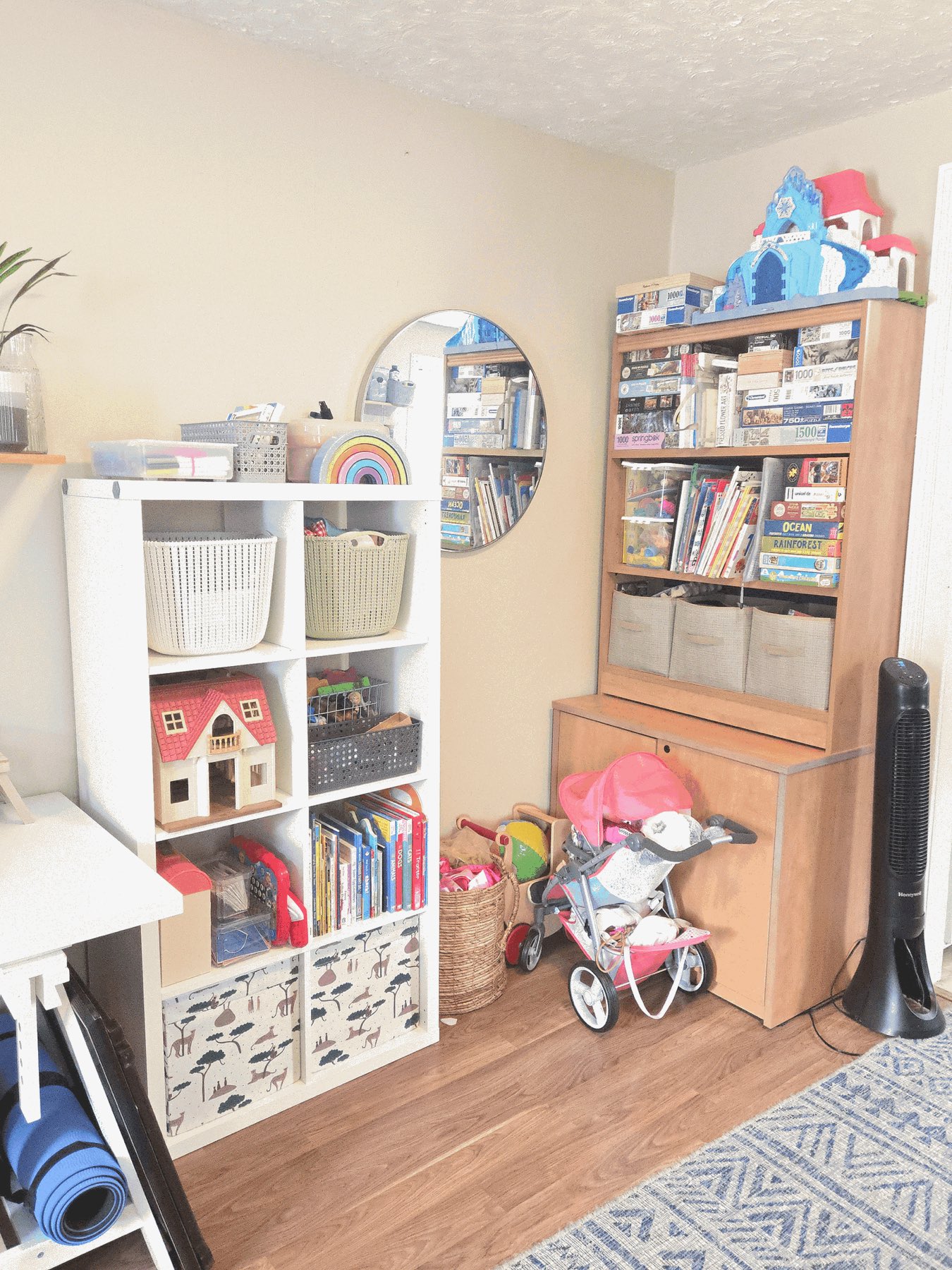 Corner of a child's playroom with a white cube shelf holding lidded baskets, a dollhouse and picture books, beside a wooden hutch of sorted puzzles and board games.